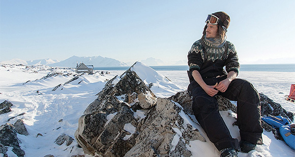 man sitting on a rock in a snowy landscape wearing ski gear with mountains and ocean in the background exploring nature in a cold environment